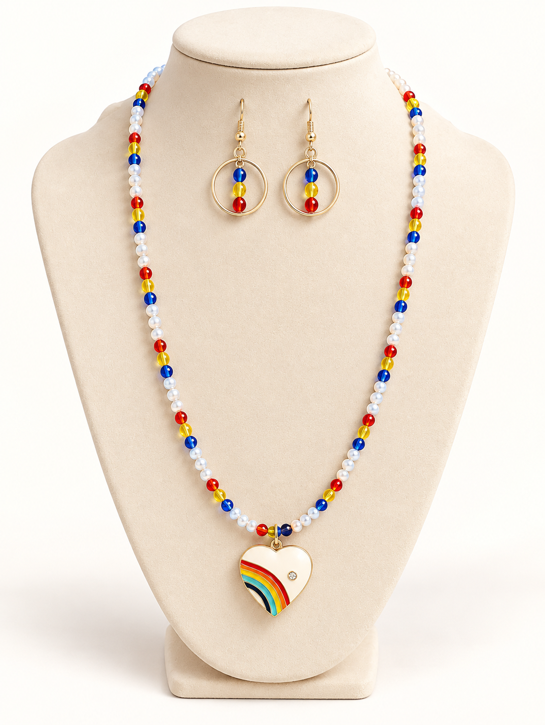 Pearl-look beaded necklace with red, yellow, and blue beads and an enamel rainbow heart pendant, with gold-tone hoop drop earrings on a cream bust